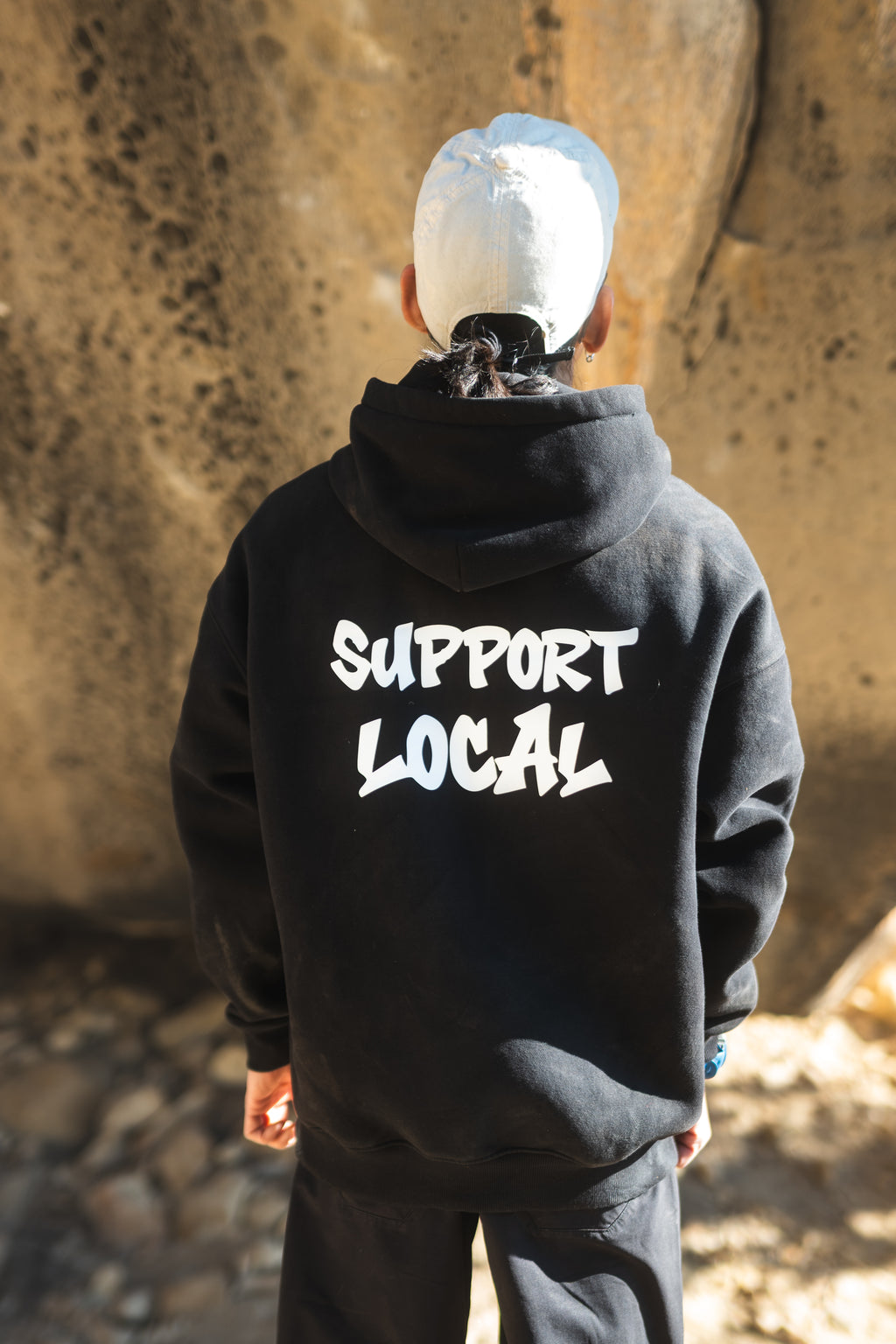 Person wearing a black hoodie with 'Support Local' text, standing against a stone wall.
