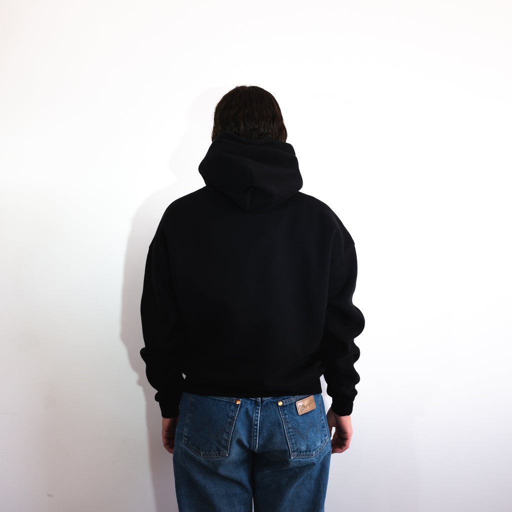 Person wearing a black hoodie and blue jeans standing against a white wall
